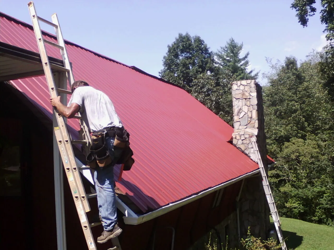Roofer on a ladder beside a red metal roof and stone chimney — Soffit & Fascia Repair by Roofing Brothers in North Weymouth